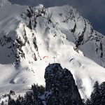 Dramatic view of paragliders soaring above snow-covered mountains and rocky peaks.