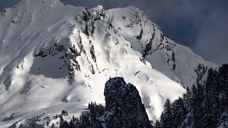 Dramatic view of paragliders soaring above snow-covered mountains and rocky peaks.