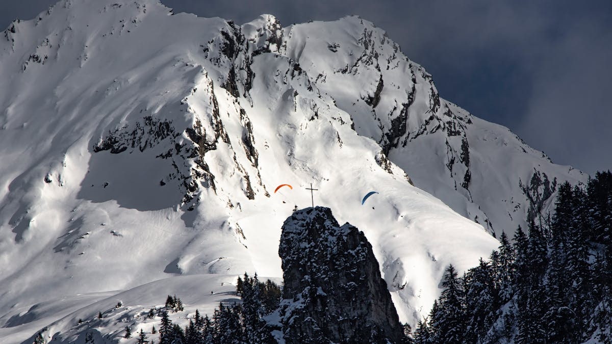 Dramatic view of paragliders soaring above snow-covered mountains and rocky peaks.